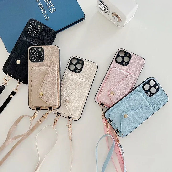 Five smartphones with pebble-texture material wallet-style cases in black, beige, pink, and blue are arranged on a white surface. Each case features a wrist strap and a cardholder pocket. A blue notebook and a small white device are also visible.