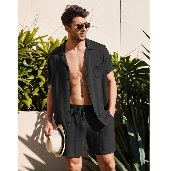 Man in black short sleeve shirt and shorts, holding hat, sunglasses, tropical background, Rovelli fashion