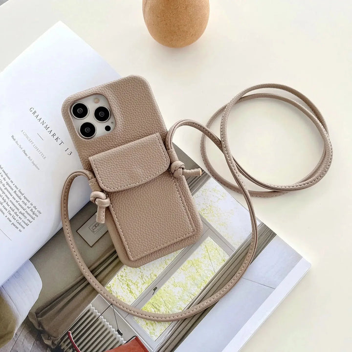 A beige leather phone case with a card holder and a long strap is placed on an open magazine and a cream-colored table. An egg-shaped object is also visible in the background.
