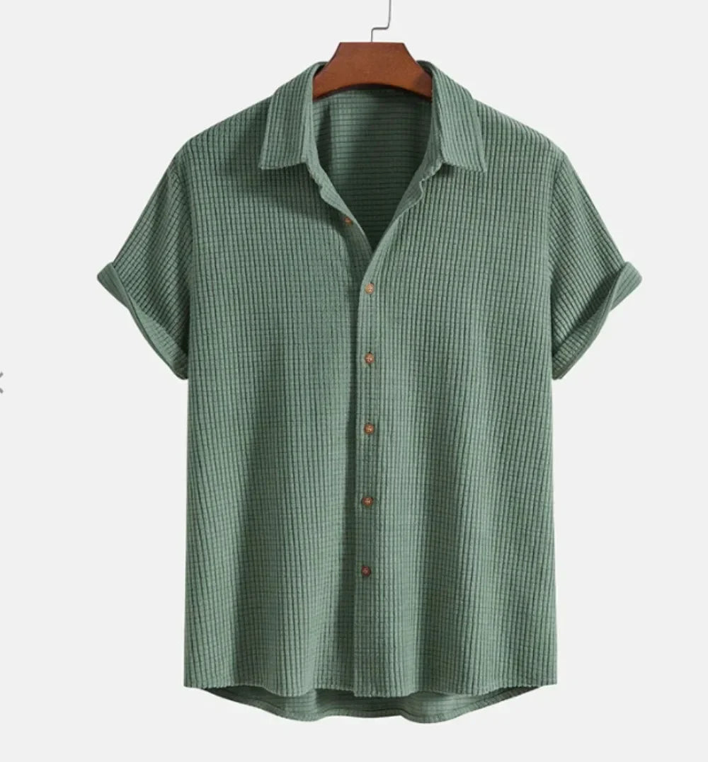 A short-sleeve, button-up Bayside Shirt in a textured, light green breathable cotton blend hangs on a wooden hanger against a plain white background. The shirt features a collar and relaxed fit—a versatile summer staple.
