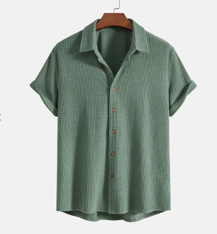 A short-sleeve, button-up Bayside Shirt in a textured, light green breathable cotton blend hangs on a wooden hanger against a plain white background. The shirt features a collar and relaxed fit—a versatile summer staple.