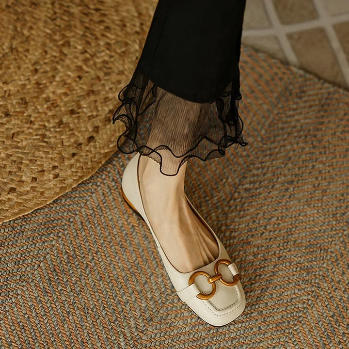 A person wearing black pants with sheer ruffled cuffs and cream-colored loafers featuring gold buckle details and a cushioned insole stands on a textured, woven rug.