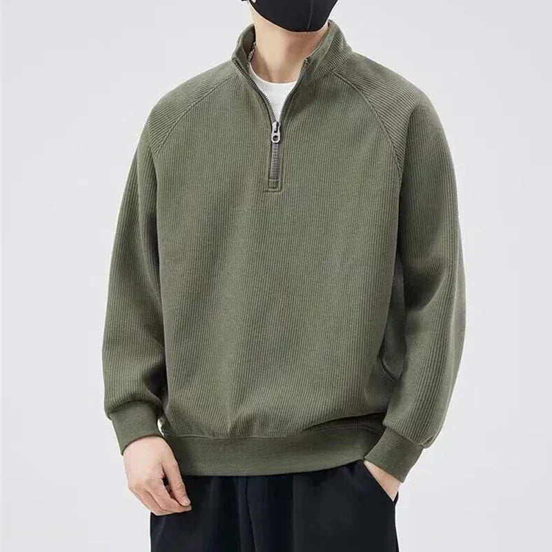 A person wearing a dark green quarter-zip pullover with a relaxed fit, black pants, and a black face mask stands against a plain light background.