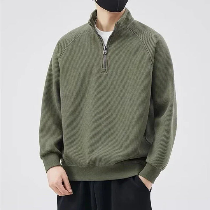 A person wearing a dark green quarter-zip pullover with a relaxed fit, black pants, and a black face mask stands against a plain light background.
