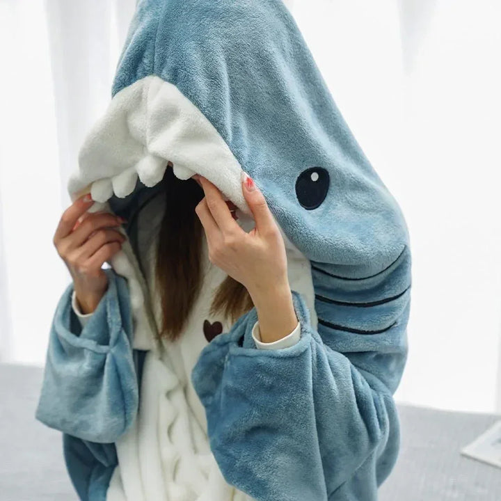 A person wearing a blue and white shark-themed hoodie with the hood pulled over their face, showing shark teeth and eyes. Only their hands and some hair are visible, giving the cozy look of shark pajamas.