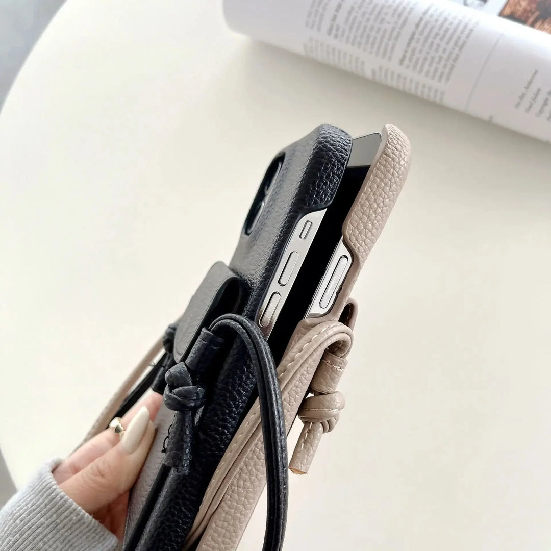 A hand holds two smartphones in textured leather phone cases—one black, one beige—each featuring a detachable lanyard with knots. Card holders are subtly integrated, while a magazine rests blurred in the background on a light-colored surface.