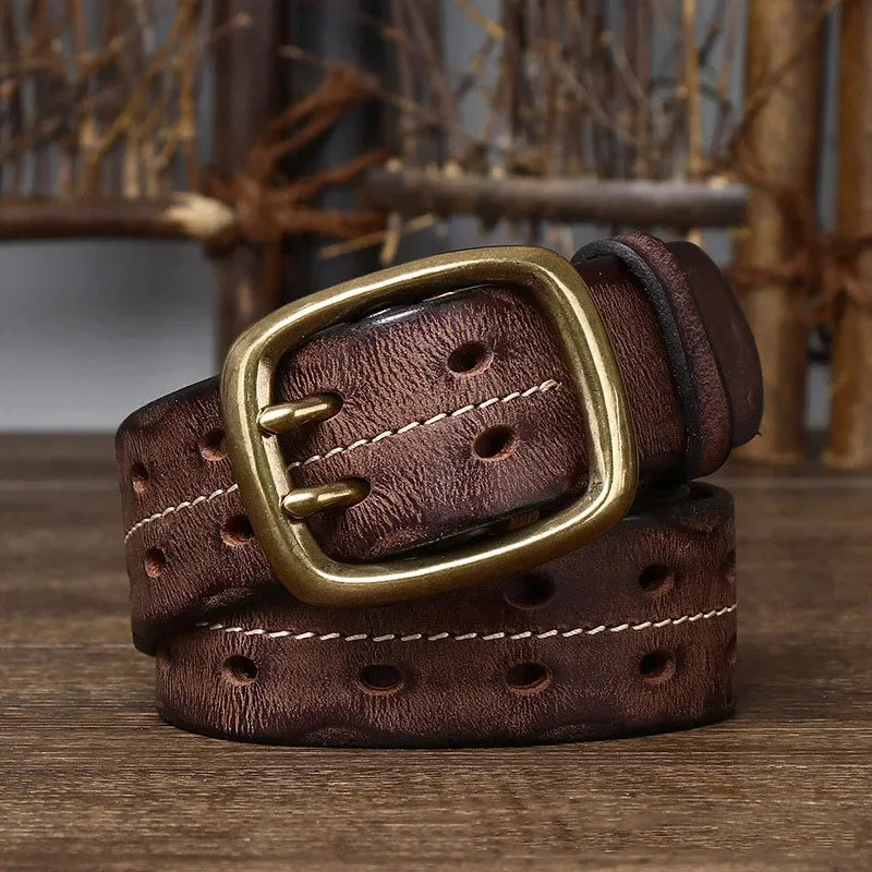 A rolled-up genuine leather belt with oval holes and a large, rectangular copper buckle sits on a wooden surface with rustic wooden sticks in the background, reflecting heritage craftsmanship.