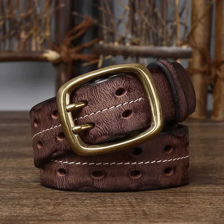 A rolled-up genuine leather belt with oval holes and a large, rectangular copper buckle sits on a wooden surface with rustic wooden sticks in the background, reflecting heritage craftsmanship.