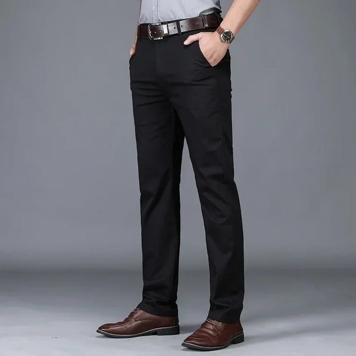 A person wearing black dress pants with a tailored fit, a light grey shirt, a brown belt, brown dress shoes, and a wristwatch, standing with hands in pockets against a plain grey background.