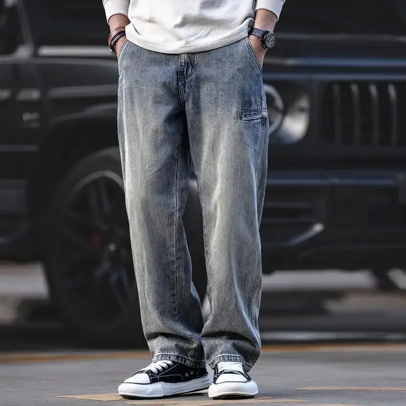 A person wearing loose-fitting, stone washed gray jeans, a white long-sleeve shirt, black wristwatch, and black-and-white sneakers stands in front of a black vehicle with hands in pockets.