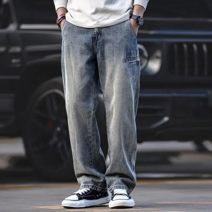 A person wearing loose-fitting, stone washed gray jeans, a white long-sleeve shirt, black wristwatch, and black-and-white sneakers stands in front of a black vehicle with hands in pockets.