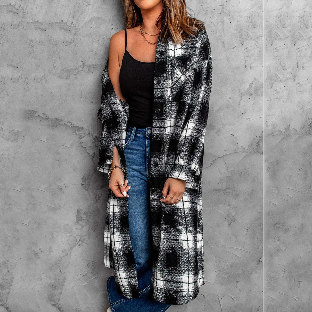 A woman stands against a gray textured wall, layering a black and white plaid long coat over a black top and blue jeans. Her coat, reminiscent of a lightweight flannel button-down shirt, is draped off one shoulder. Her wavy brown hair hides her face.