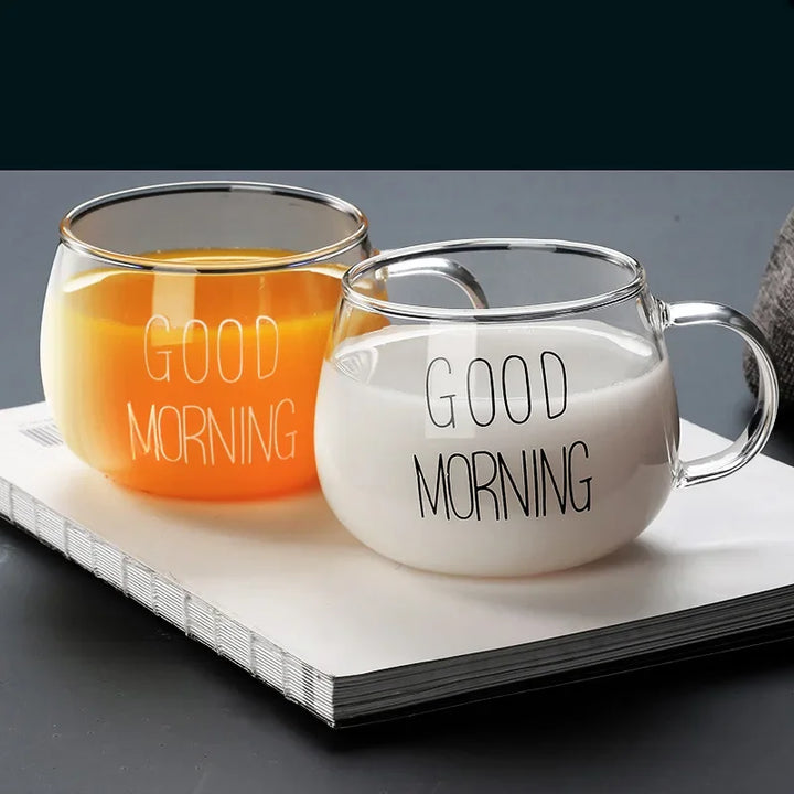 Two clear glass mugs featuring a clear design and “GOOD MORNING” print, one with orange juice and the other with milk, rest side by side on a closed white book against a dark background.