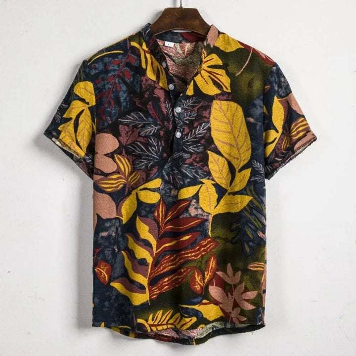 A short-sleeved Vintage Shirt Men with an oversized fit, featuring a tropical leaf pattern in yellow, red, orange, and pink on a dark background, hanging on a wooden hanger against a plain white wall—perfect for summer streetwear.
