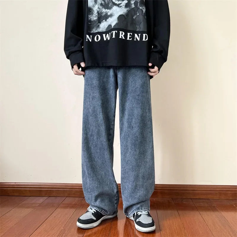 Person standing indoors on a wooden floor, wearing relaxed fit blue jeans in a vintage wash, black and white sneakers, and a black sweatshirt with NOWTREND written on it. The persons face is not visible.