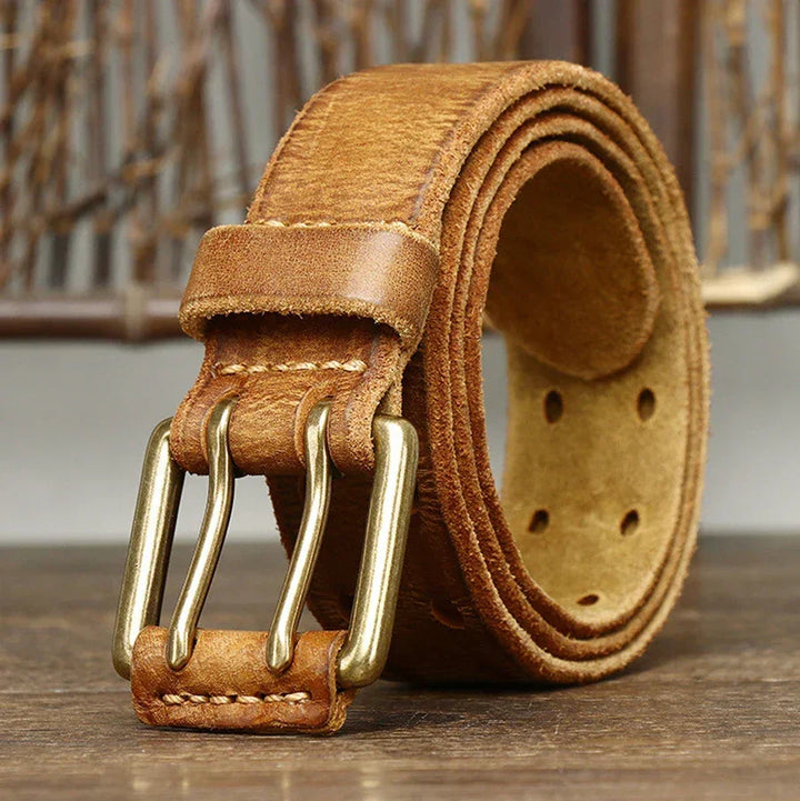 A rolled-up genuine leather belt with a double brass buckle rests on a wooden surface, showcasing heritage craftsmanship against a blurred rustic background.