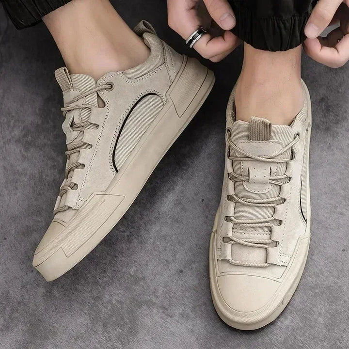 A person wearing beige lace-up sneakers with a cushioned insole and thick soles stands on a gray surface. One foot is flat while the other is slightly raised as they adjust the laces, highlighting the shoes’ textured design.