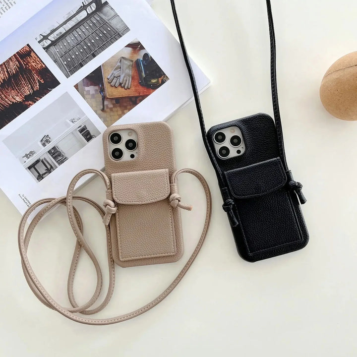 Two smartphones in leather phone cases, one beige and one black, lie on a white surface beside a magazine and a brown spherical object. Each case features small pouches with card holders and long detachable lanyards for hands-free convenience.