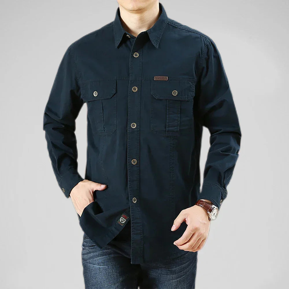 A man wearing a relaxed fit dark blue button-up shirt with two front pockets, dark jeans, and a wristwatch stands with one hand in his pocket against a plain light gray background.