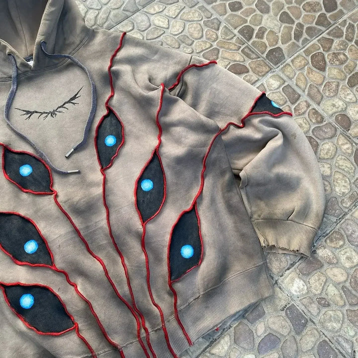 A tan hooded sweatshirt with a comfortable loose fit and red-stitched, abstract eye pattern patchwork featuring blue and black centers, displayed on a stone-tiled surface. The hem and sleeve cuffs are frayed for a distressed look.