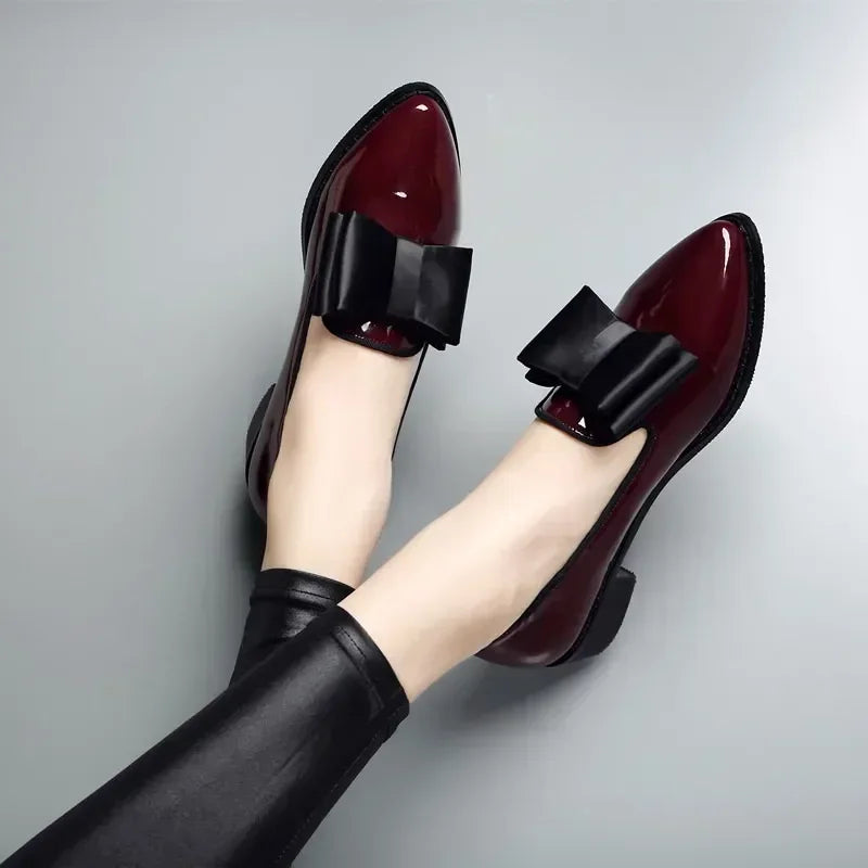 A person wearing shiny patent leather burgundy loafers with large statement bows and black leggings, resting their feet on a light gray surface.
