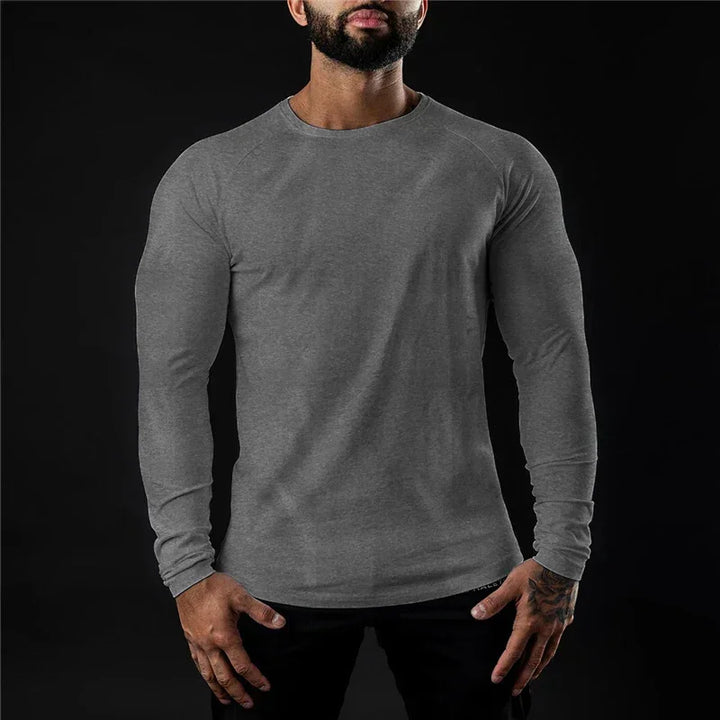 A man wearing a long sleeve fitted shirt stands against a black background. His face is partially out of frame, and his arms are relaxed at his sides.