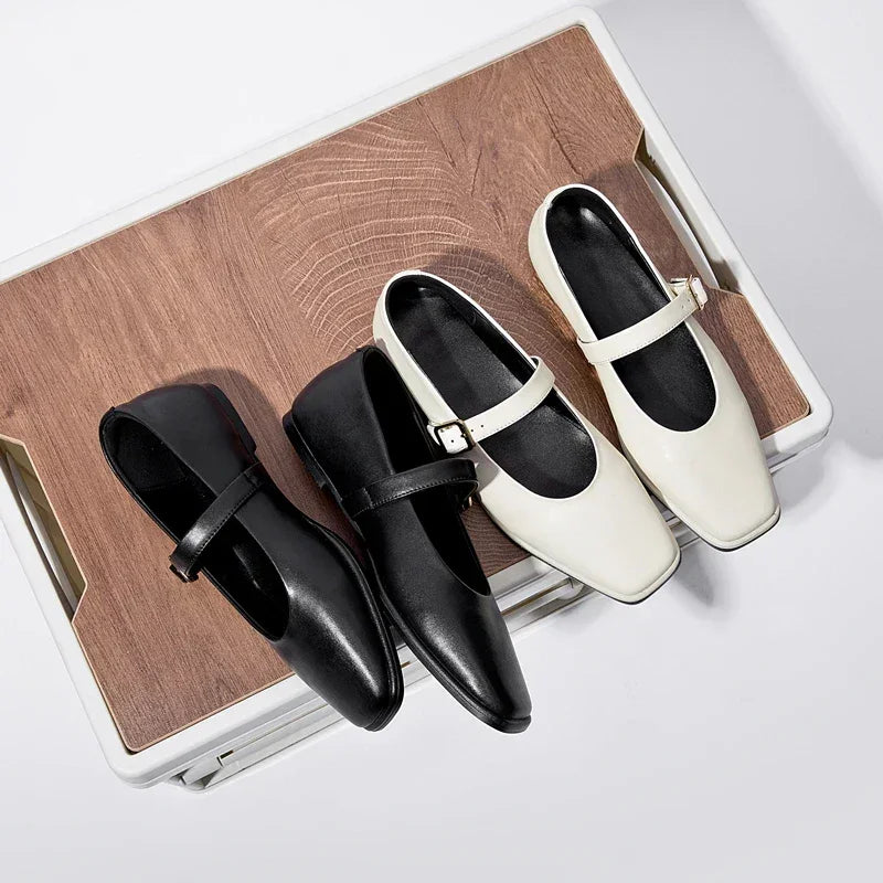 Two pairs of Mary Jane shoes, one black and one white, with rounded toes and single straps, are displayed side by side on a wooden surface with a light geometric pattern, resembling classic ballet flats for a timeless look.