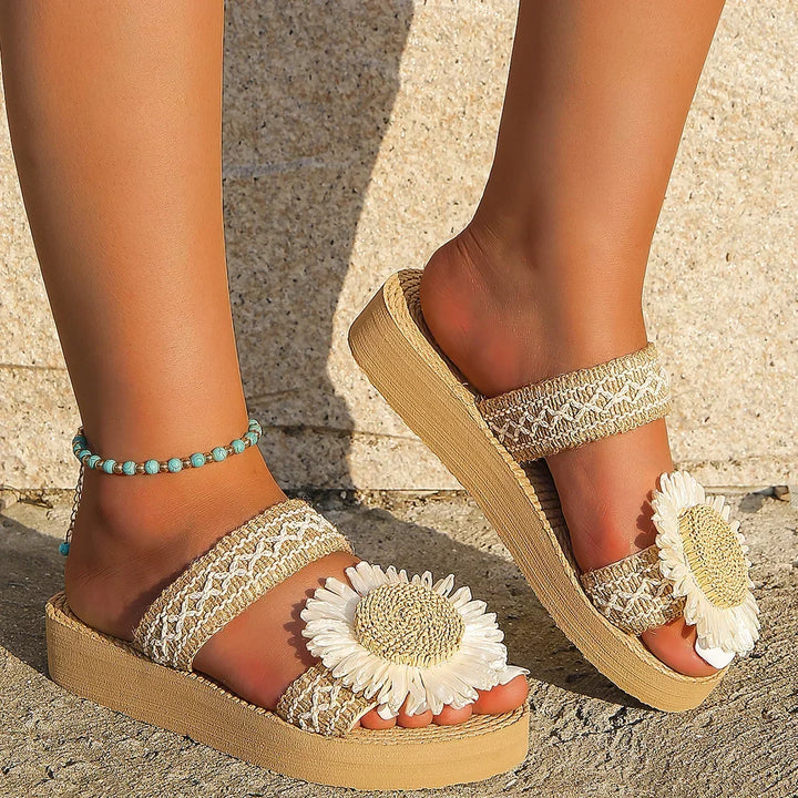 A pair of woven women's fashion sandals with large white daisy flowers on the straps are worn by someone standing on sandy ground, accented by a turquoise beaded anklet—perfect summer footwear.