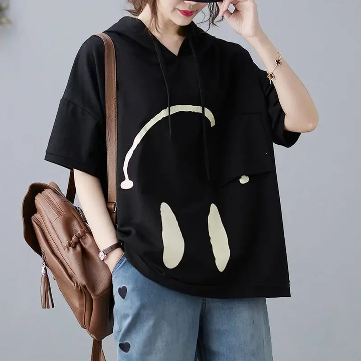 A woman wears a black Monday Mood hoodie with a large, abstract yellow smiley face, blue jeans with heart-shaped patches, and a brown backpack over one shoulder. She touches her face and smiles slightly, ready for spring colors.