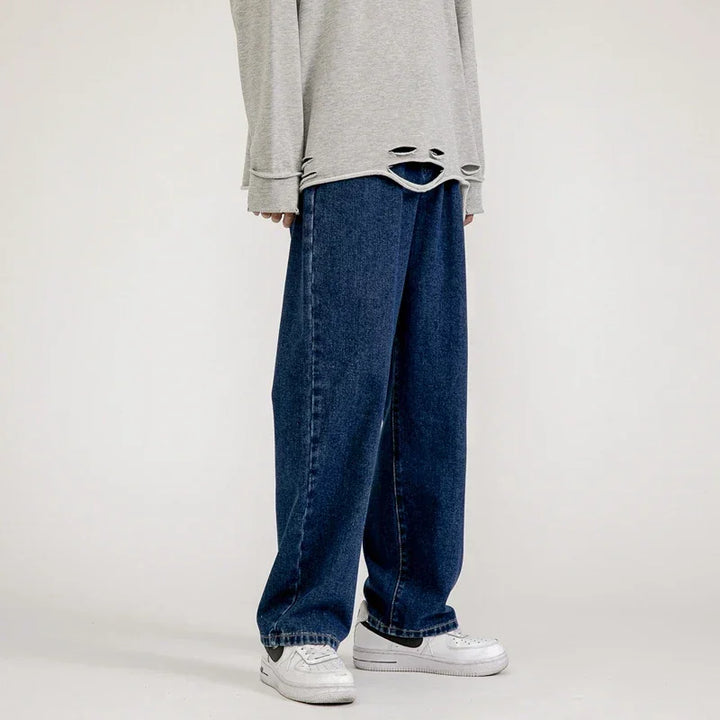 Person wearing a loose, light gray sweater with distressed holes at the hem, relaxed fit jeans in a dark vintage wash, and white sneakers, standing against a plain background.