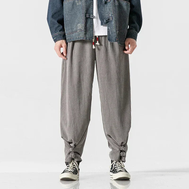 A person wearing loose, gray cotton blend sweatpants with a drawstring waistband tied at the ankles, black and white sneakers, a white shirt, and a blue denim jacket, standing against a plain light background.