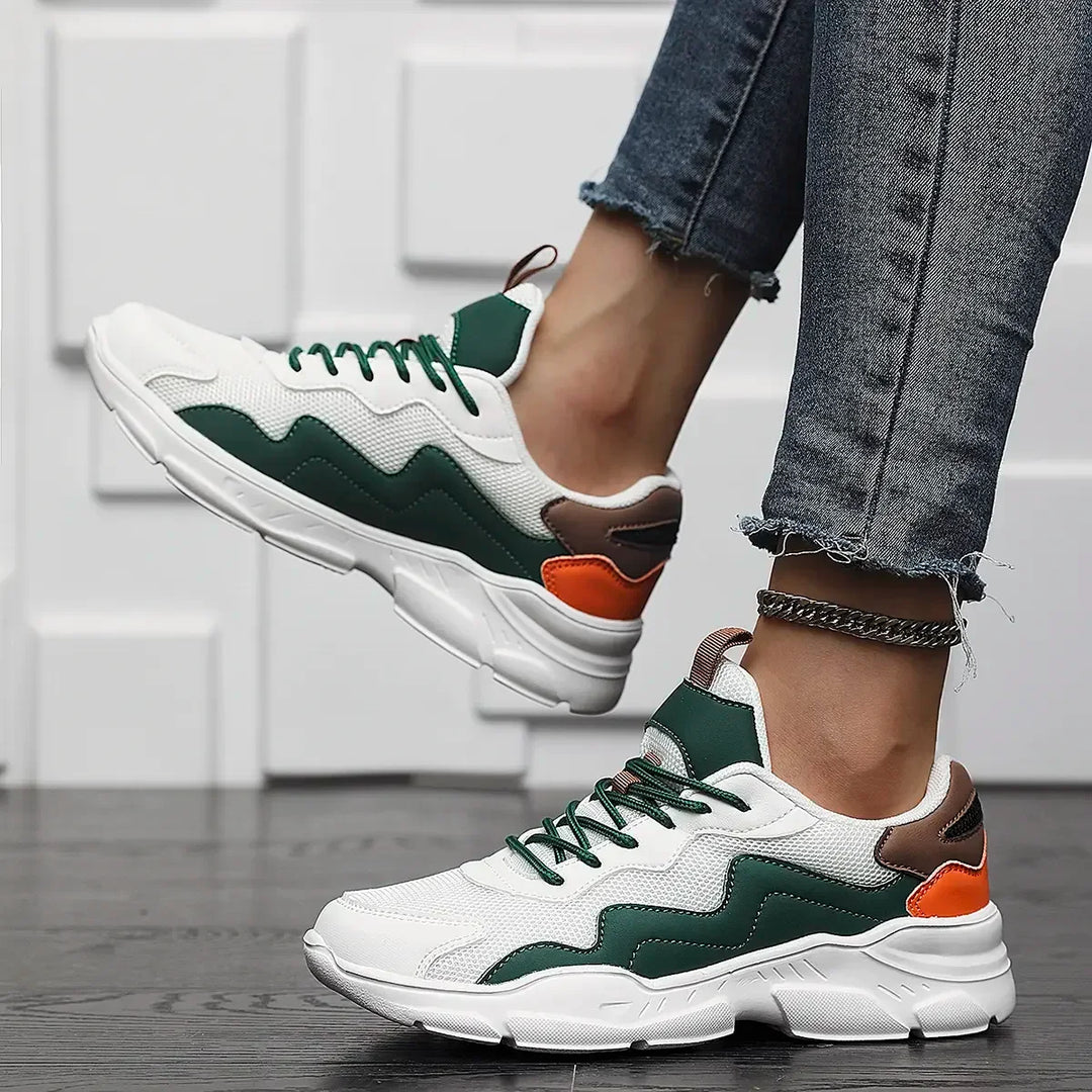 A person wearing advanced shock absorption sneakers in white, green, brown, and orange stands on dark wooden flooring. They are dressed in frayed-hem blue jeans and an anklet, with their feet lifted slightly off the ground.