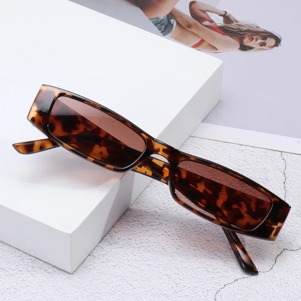 Retro rectangle sunglasses with UV400 protection and brown lenses rest on a white surface, complemented by a partial photo of a woman in a bikini visible in the background.