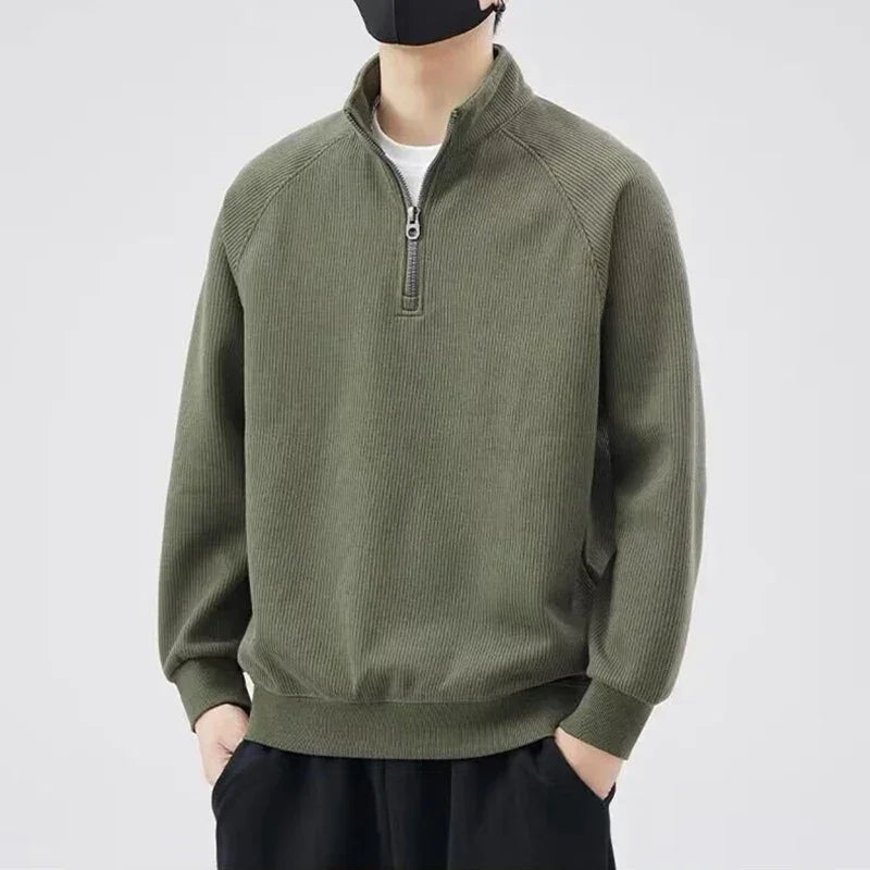 Person wearing a dark green ribbed quarter-zip pullover sweater with stand-up collar, hands in pockets, black pants, and a black face mask, standing against a plain light background.