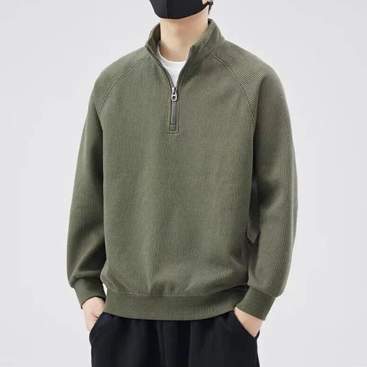 Person wearing a dark green ribbed quarter-zip pullover sweater with stand-up collar, hands in pockets, black pants, and a black face mask, standing against a plain light background.