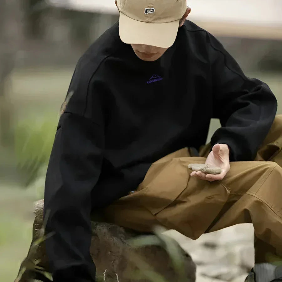Man in black sweatshirt and khaki pants sitting outdoors, wearing beige cap, Rovelli fashion