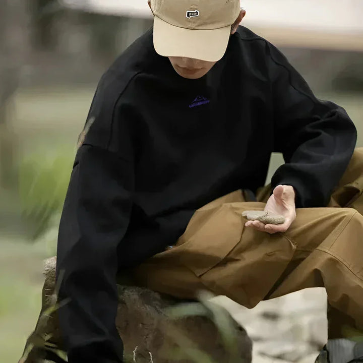 Man in black sweatshirt and khaki pants sitting outdoors, wearing beige cap, Rovelli fashion