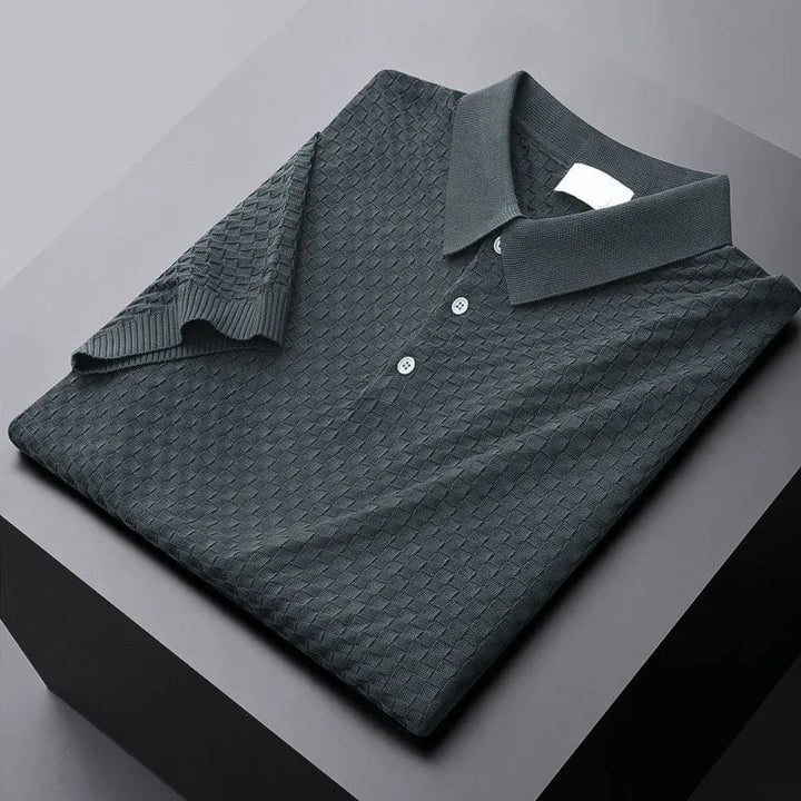 A neatly folded dark gray polo shirt with a textured, diamond waffle jacquard pattern and three buttons, placed on a black and gray surface.