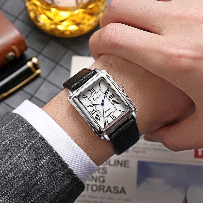 A person in a gray pinstripe suit wears a luxury watch with a square silver case, white dial, black Roman numerals, and premium leather band. A glass of whiskey, pen, and newspaper rest in the background.