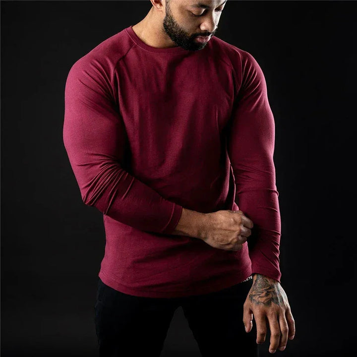 A man wearing a long sleeve fitted, moisture-wicking shirt and black pants stands against a dark background, looking down and adjusting his sleeve with one hand.