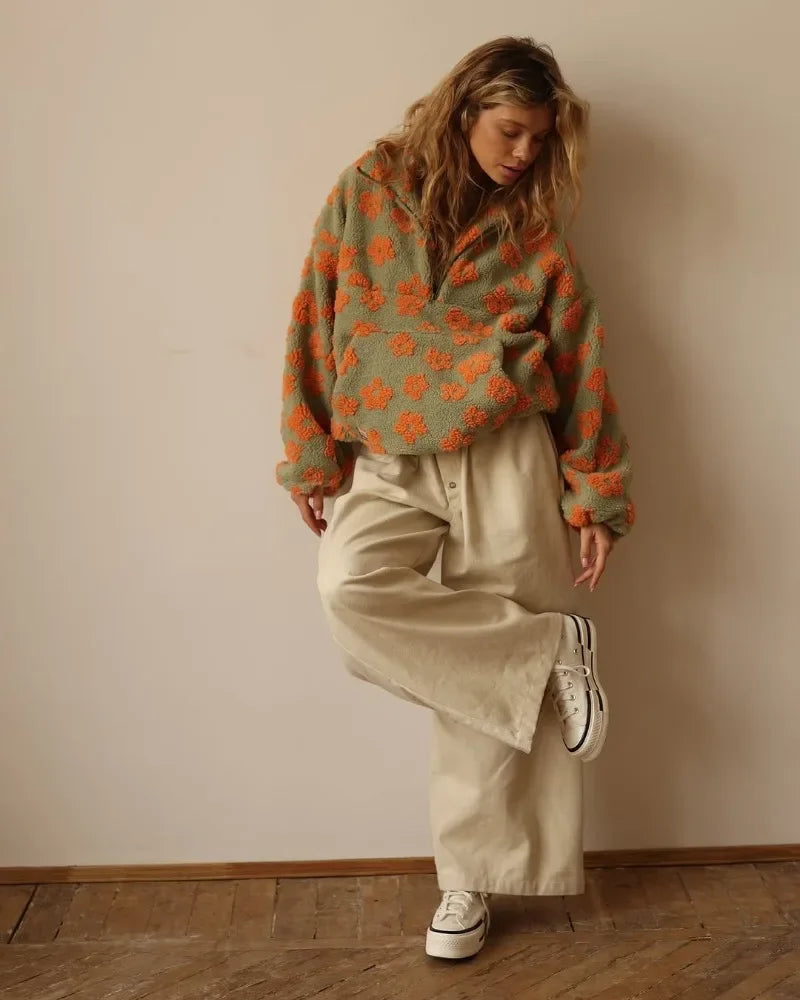A person wearing an office lady sweater with orange flower patterns, wide beige pants, and white sneakers stands indoors on a wooden floor, balancing on one foot and looking down.