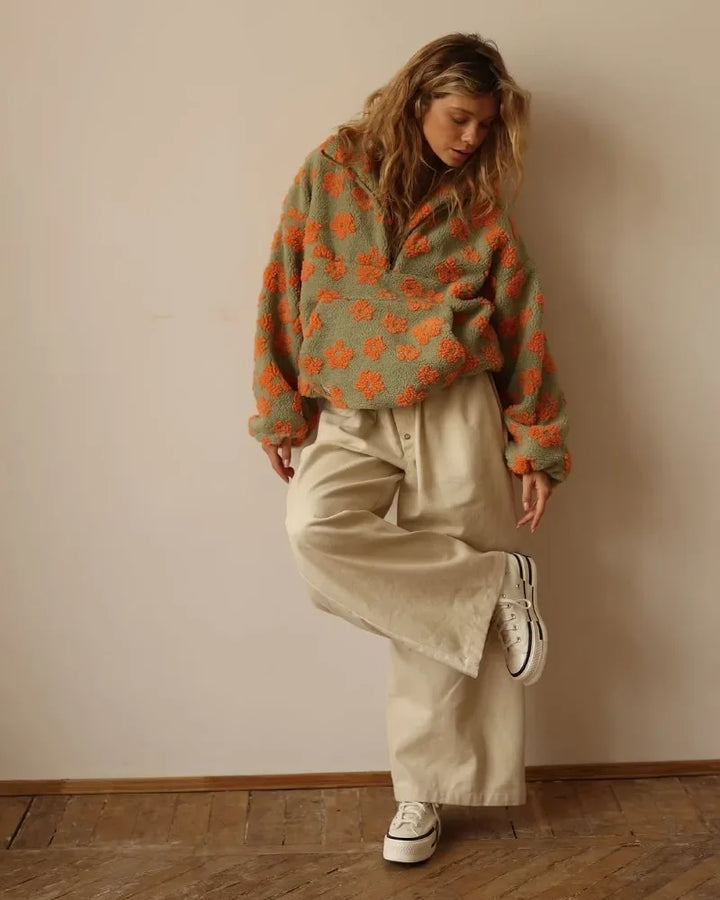 A person wearing an office lady sweater with orange flower patterns, wide beige pants, and white sneakers stands indoors on a wooden floor, balancing on one foot and looking down.
