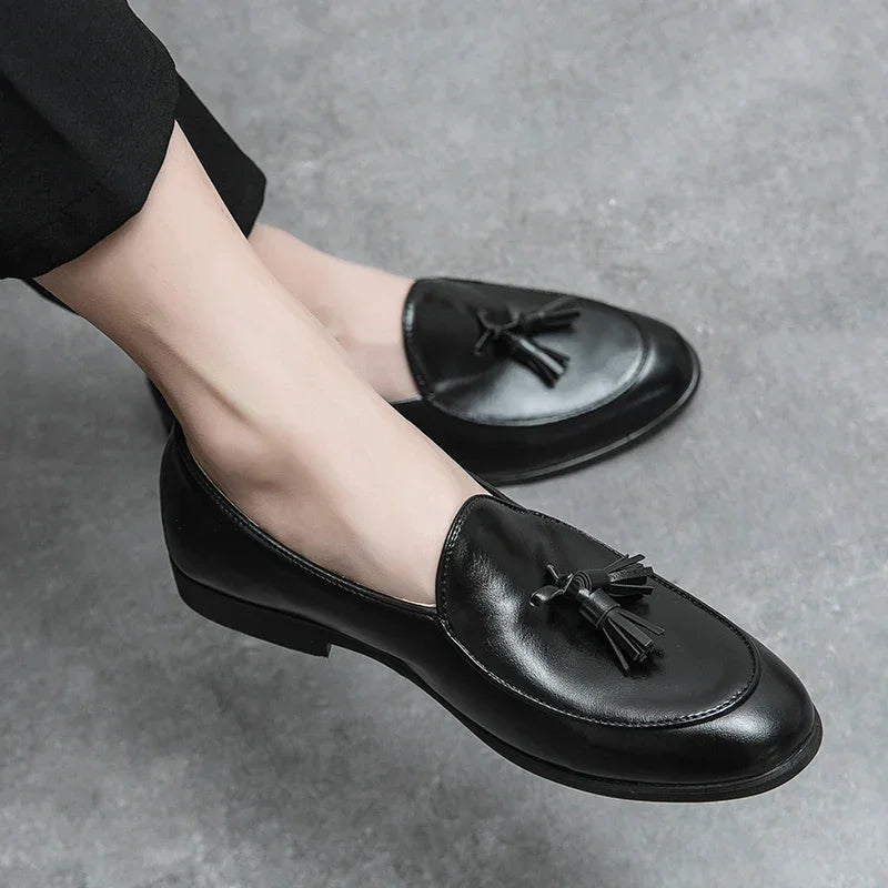 A person wearing black leather tassel loafers with a cushioned insole and black pants, seated on a gray textured surface.