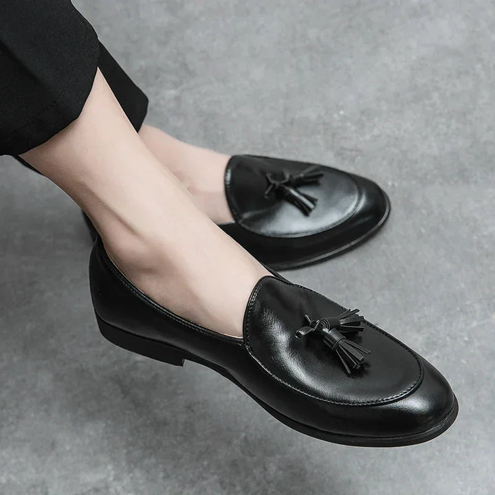 A person wearing black leather tassel loafers with a cushioned insole and black pants, seated on a gray textured surface.