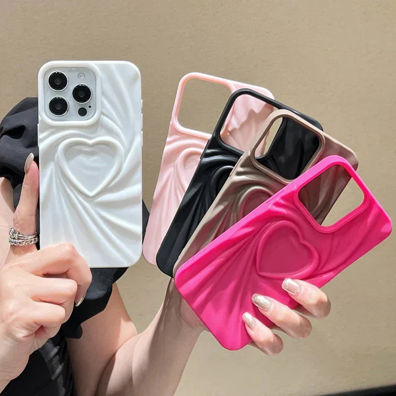 A person holds five phone cases in soft matte silicone with 3D heart designs. Featuring full coverage protection, the cases come in white, light pink, black, fuchsia, and beige against a neutral background.