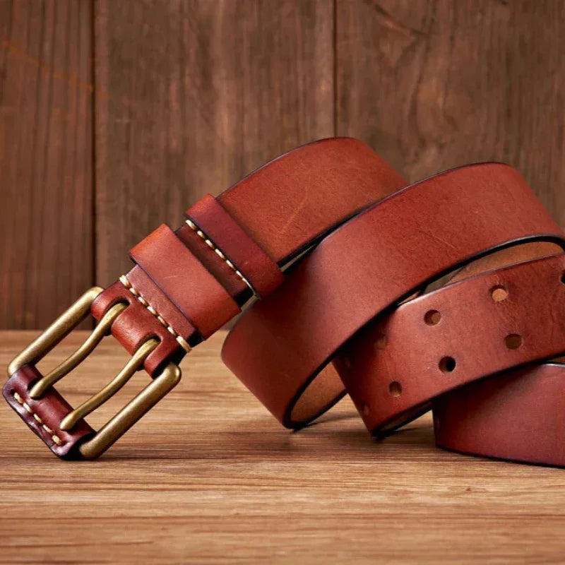 A brown cowhide leather belt with a copper buckle is coiled on a wooden surface, set against a rustic background. The durable belt features visible stitching near the buckle and multiple adjustment holes.