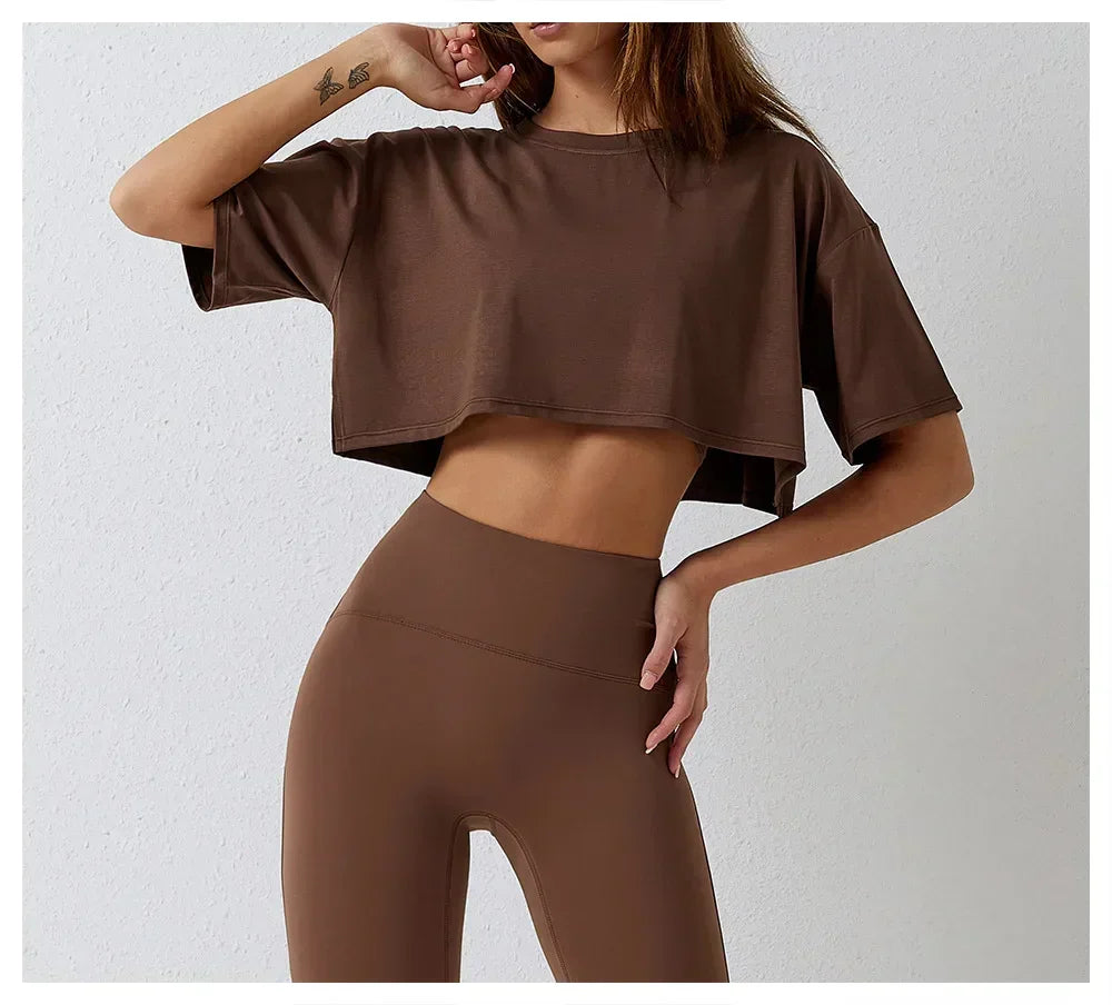 A woman poses against a light background, wearing a cotton crop top and matching high-rise pants. Her face is partially out of frame, with one hand on her hip and the other touching her chin.