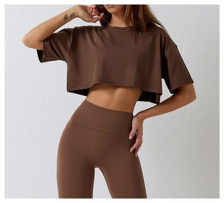 A woman poses against a light background, wearing a cotton crop top and matching high-rise pants. Her face is partially out of frame, with one hand on her hip and the other touching her chin.