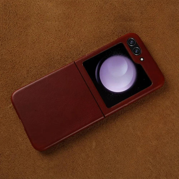 A Samsung Galaxy Z Flip with a genuine leather case rests on a textured brown surface. The screen displays a large white and purple sphere, while dual camera lenses add style and shockproof protection in the top right corner.