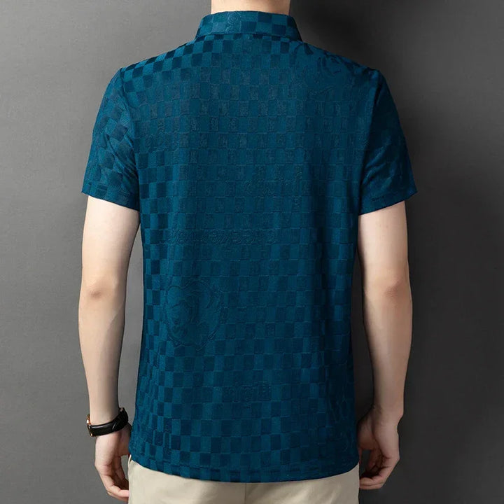 A person wearing a short-sleeve, teal checkerboard pattern polo shirt and beige pants with a tailored fit is standing with their back to the camera against a neutral background.